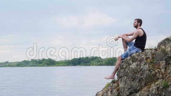 年轻人坐在一个乡村湖泊上方的岩石悬崖上乡村男孩孤独的年轻人视频的预览图