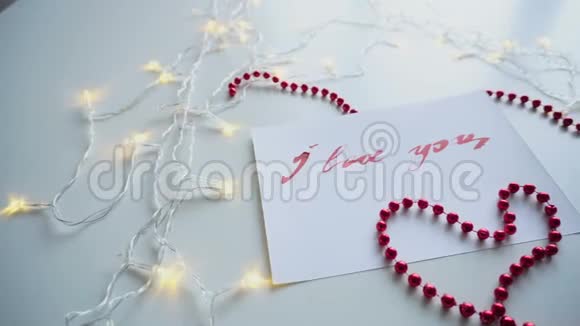 漂亮的书法卡片红色的铭文我爱你在白色的纸上桌子上闪烁的花环和珠子视频的预览图