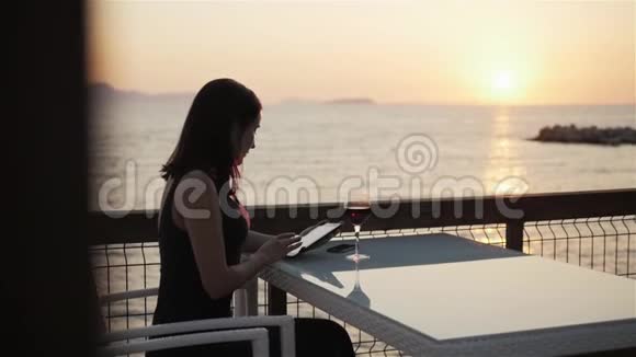 日落时分坐在海边的咖啡馆里年轻的布鲁内特用她的平板电脑端着一杯红酒视频的预览图