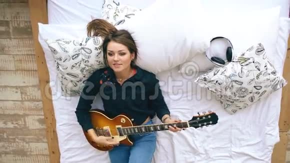 有魅力的年轻女孩在家里的卧室里躺在床上学弹电吉他视频的预览图
