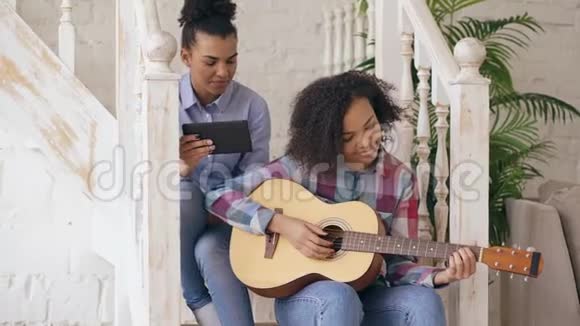 混合种族的年轻女子坐在楼梯上教她十几岁的妹妹在家弹吉他视频的预览图