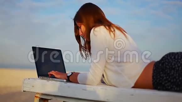 女人躺在海边的长凳上在笔记本电脑上打字视频的预览图