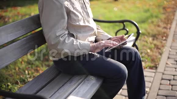 英俊的老人坐在长凳上用平板电脑在互联网上滚动秋树的背景视频的预览图