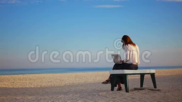 白种人女孩在坐在沙滩上的长椅上的笔记本电脑上写着文字沿着海滨的泡泡视频的预览图
