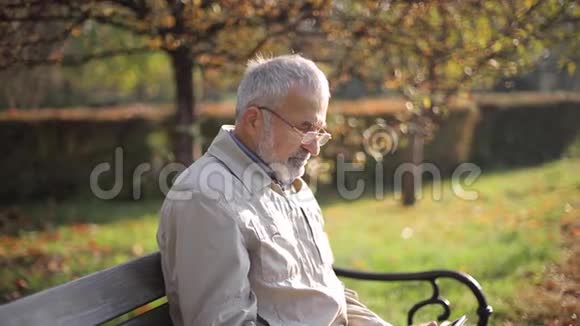 英俊的老人坐在长凳上用平板电脑在互联网上滚动秋树的背景视频的预览图