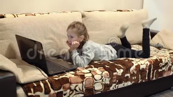 有趣的小女孩坐在沙发上用笔记本电脑和微笑视频的预览图