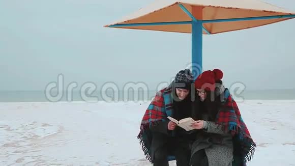 一个女孩和一个男人坐在海滩的天篷下上面铺着一个格子在看书视频的预览图