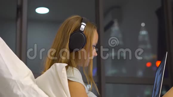 年轻女子戴着无线耳机用笔记本电脑工作她坐在床上一个摩天大楼的剪影视频的预览图