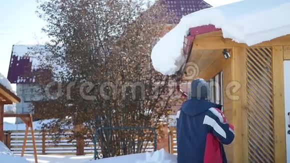 一个穿着冬季夹克的年轻人用塑料铲子屋顶脱掉了很多雪站在地上下面的动作视频的预览图