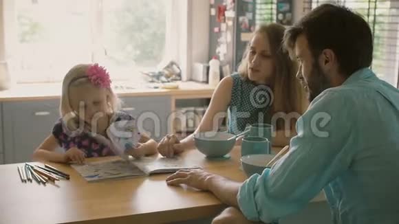 当她的父母在厨房的餐桌上吃早餐时小女孩正在给她的父母看她的彩色书慢节奏视频的预览图