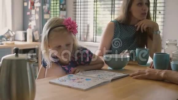 可爱的金发小女孩看着她的着色书而父母在厨房的桌子上交谈慢点斯泰迪达姆视频的预览图