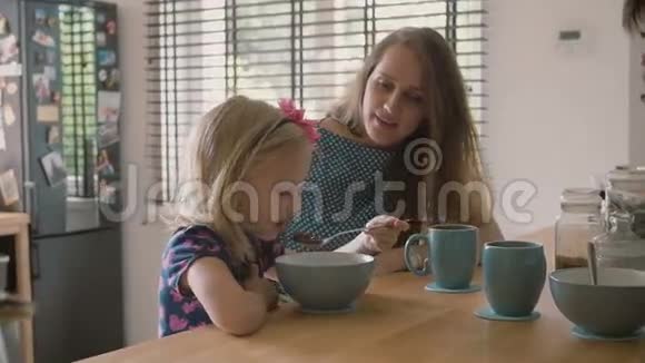 美丽的年轻母亲和她可爱的金发女儿在厨房的餐桌上快乐地笑着慢节奏视频的预览图
