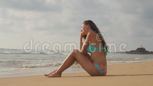 美丽的年轻女子比基尼坐在沙滩上的金色沙滩上在完美的天堂海岸放松的女孩视频的预览图