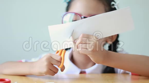 穿着泰国幼儿园校服的亚洲小女孩用剪刀在木桌上剪白纸视频的预览图
