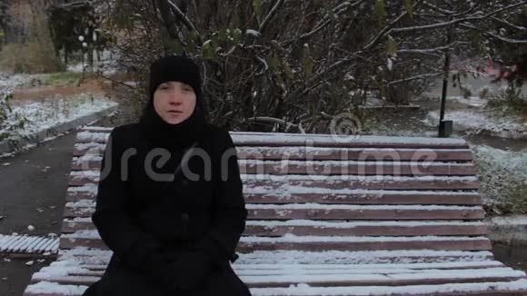 冬天一个穿着黑裙子的女孩坐在公园的长凳上的肖像视频的预览图