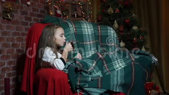 女孩喝茶在圣诞树附近的椅子上玩视频的预览图