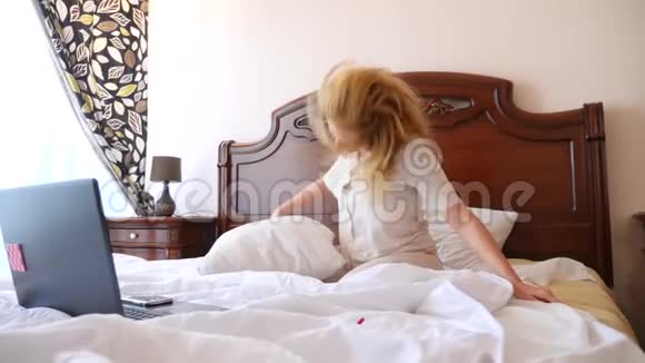 穿着睡衣的女人从笔记本电脑传来的欢乐的消息在床上欢快地跳舞4k慢动作视频的预览图