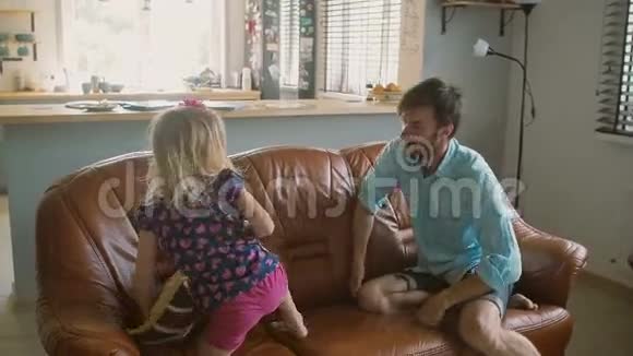 年轻的爸爸在沙发上和他可爱的女儿玩枕头和她打架挠痒痒慢点斯泰迪达姆视频的预览图