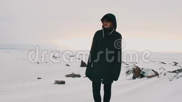 年轻的金发女郎穿着黑色夹克和帽子站在冰冻的湖岸上视频的预览图
