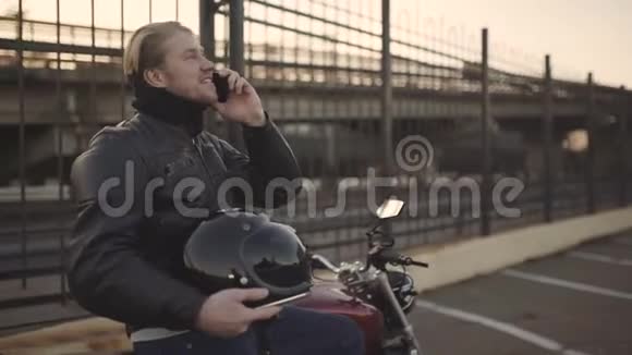日落时分一个身穿皮夹克的年轻人坐在摩托车上打电话视频的预览图