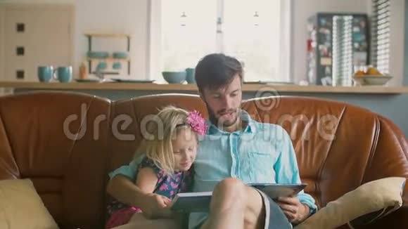 一位年轻的父亲和他快乐的金发小女儿正在餐厅的皮革沙发上读一个故事慢动作视频的预览图