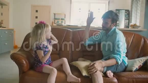 一位年轻的父亲和他可爱快乐的小女儿正坐在沙发上互相给予对方高昂的生活慢动作视频的预览图