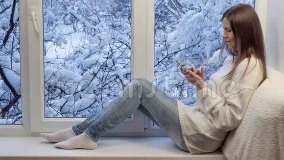 漂亮的女孩坐在窗台上看着窗户用平板电脑冬天在外面视频的预览图