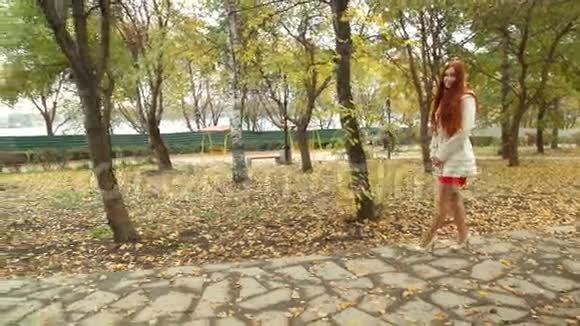 穿着白色大衣的女孩留着红色的长发扫着秋公园的林荫道视频的预览图