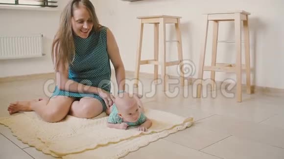 美丽的母亲正微笑着看着她的孩子试图爬在厨房地板上的毯子上慢节奏视频的预览图