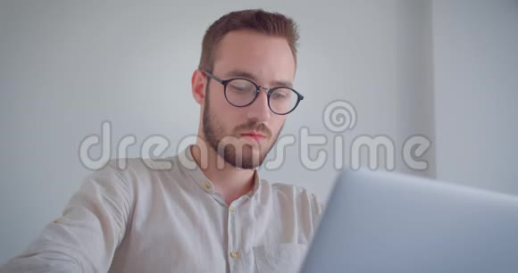 年轻英俊的白种人商人在笔记本电脑上打字在室内看着照相机的特写照片视频的预览图