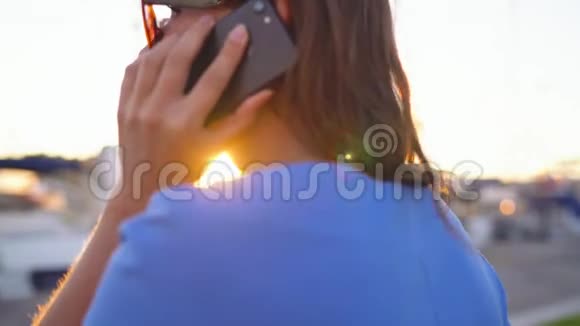 戴着太阳镜的女人在日落时分带着很多游艇和船沿着码头走着用智能手机做特写慢慢视频的预览图