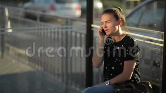 美丽的年轻女子等着公共汽车看着她的手表女孩在公共场合用手机说话视频的预览图