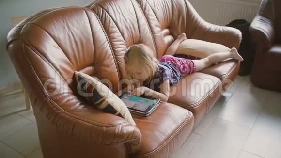 穿着粉色衣服的金发小女孩躺在沙发上在平板电脑上玩游戏慢动作视频的预览图