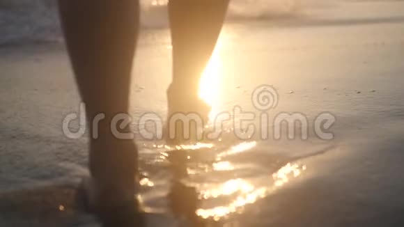 美丽的女性双脚在美丽的日落背景下带着波浪和飞溅海滨散步高清高清视频的预览图