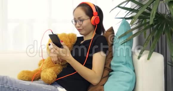 美丽的亚洲女人正在听音乐耳机在家里的沙发上使用智能手机放松生活方式视频的预览图