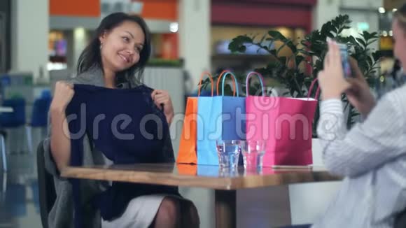 快乐的亚洲女人看着一件新衣服她的金发朋友在购物中心给她拍照视频的预览图