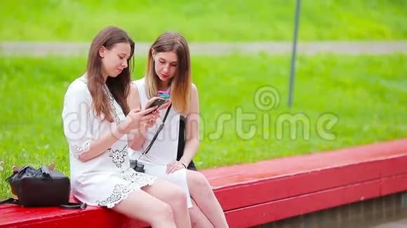 两个女孩在户外使用智能手机两个女人坐在公园里用智能手机发送信息视频的预览图