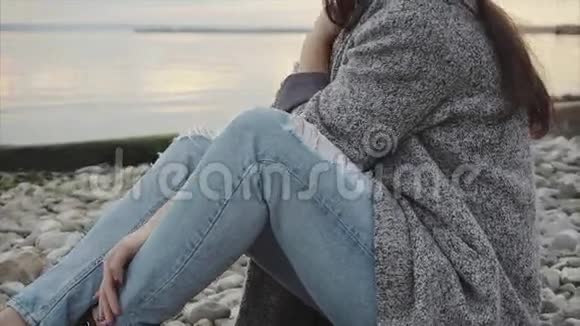 戴着黑帽子戴着眼镜的年轻漂亮微笑的女人坐在夕阳下的海边视频的预览图