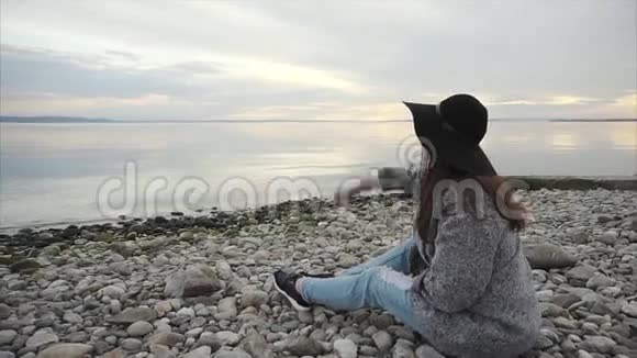 年轻女子坐在鹅卵石海滩上向水中扔石头视频的预览图