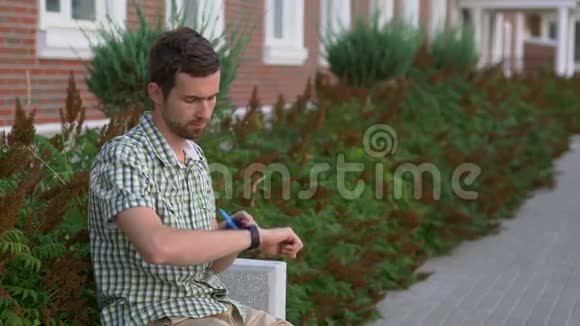 休闲打扮的男人坐在长凳上用他的智能手表和智能手机视频的预览图