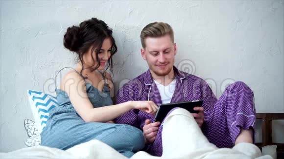 年轻男女穿着睡衣坐在床上在平板电脑上看视频视频的预览图