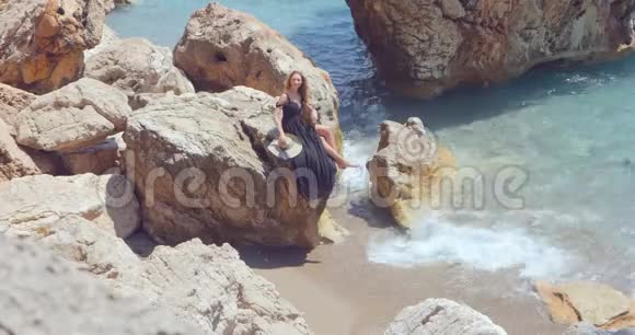 年轻漂亮的女人穿着一件黑色优雅的衣服坐在海边的岩石上视频的预览图