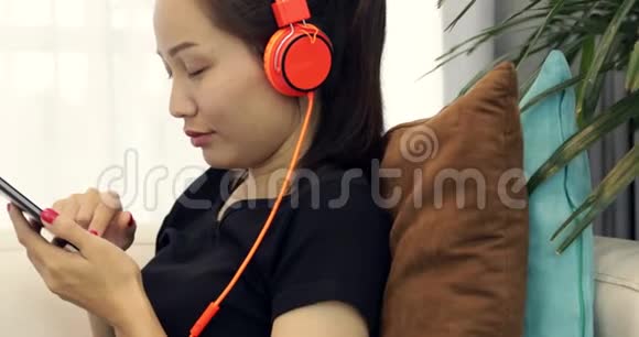 美丽的亚洲女人正在听音乐耳机在家里的沙发上使用智能手机放松生活方式视频的预览图
