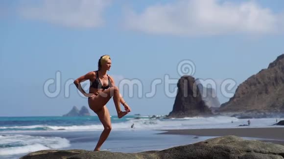 穿着比基尼站在一条腿上做瑜伽的年轻女孩站在海边的一块黑沙滩上视频的预览图