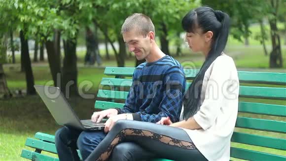 恋爱中的情侣坐在公园的长凳上看着一台笔记本电脑视频的预览图