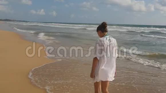 美丽的女人穿着泳衣和衬衫在海边散步举起双手年轻女孩在海边视频的预览图