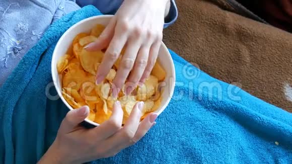 女人躺在蓝色毯子下的沙发上吃着深盘子里的薯片不健康食品快餐视频的预览图