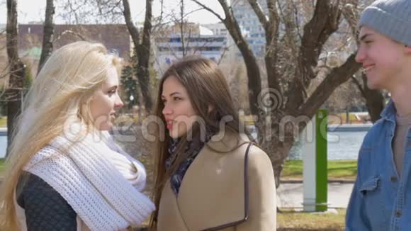 在公园里见朋友两个金发和黑发女孩站着说话一个年轻人走近他们他们兴高采烈视频的预览图