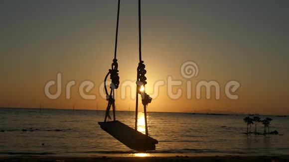 在美丽的日落和灿烂的阳光下在热带海滩海边的椰子树上挂着木制秋千椅的剪影视频的预览图