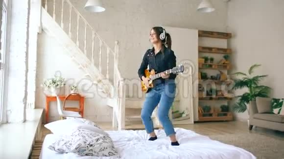 有趣的年轻女子戴着耳机在床上蹦蹦跳跳家里的卧室里放着电吉他视频的预览图